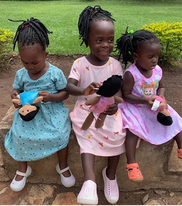 Three young girls in colorful dresses sit together, holding dolls, with a grassy background.