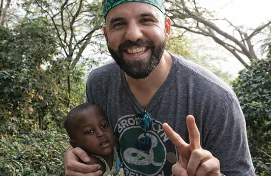 A smiling man in a green headband holds a young boy, both posing with peace signs in a natural setting.