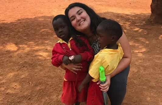 A smiling volunteer hugs two young children in colorful outfits, set against a dusty, outdoor backdrop.