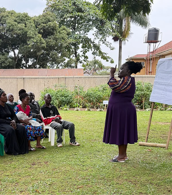 A woman speaks to a group seated outdoors in a green area, with a whiteboard and trees in the background.