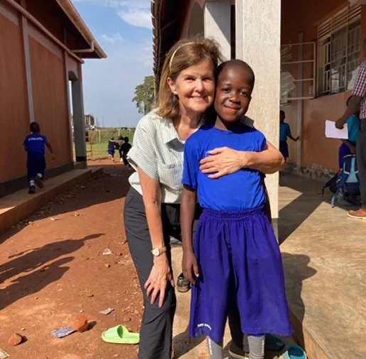 Smiling woman kneels beside a young boy, both wearing casual clothes, outdoors near a school building.