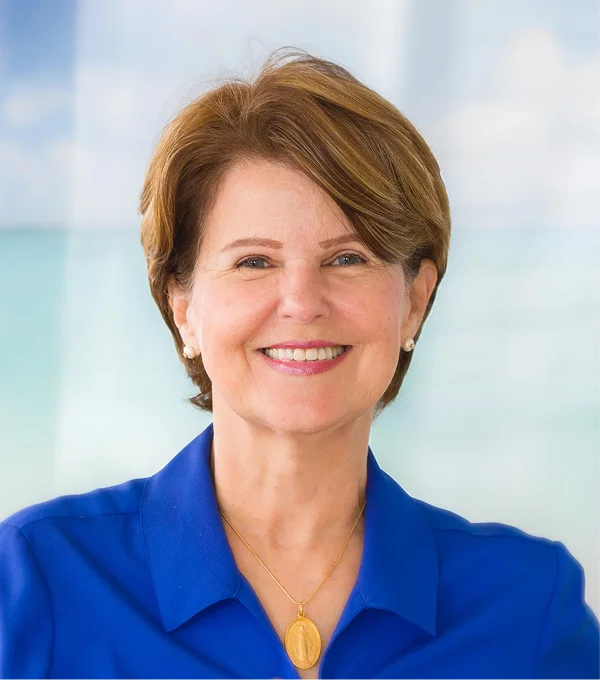 Smiling woman with short brown hair wearing a blue blouse and a necklace, set against a soft, light background.