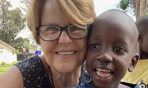 Smiling woman with glasses embraces a smiling boy outdoors, surrounded by greenery and blurred children in the background.
