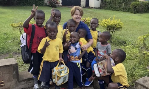 Woman surrounded by smiling children in yellow shirts, posing together outdoors with a green backdrop.