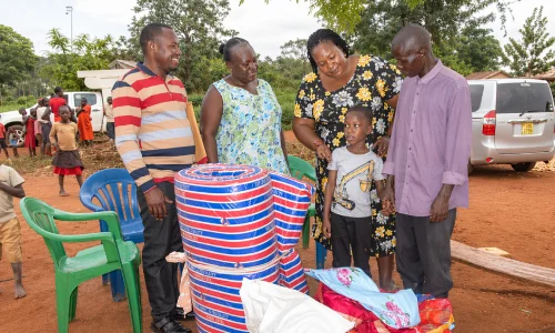 A group of five people gathers around colorful rolled goods, with children playing in the background.