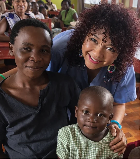 A smiling woman with curly hair sits with a child in a green outfit and another person in a classroom setting.