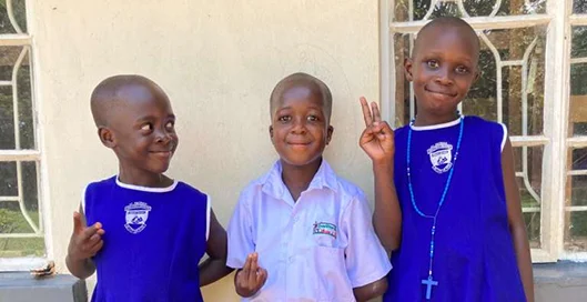 Three children pose for a photo, smiling and making peace signs, standing against a light wall.