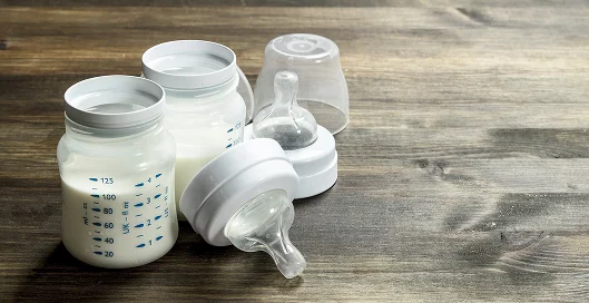Two baby bottles and two nipples filled with milk on a wooden surface.